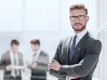 Mężczyzna w garniturze i okularach stoi z rękami skrzyżowanymi na piersi.